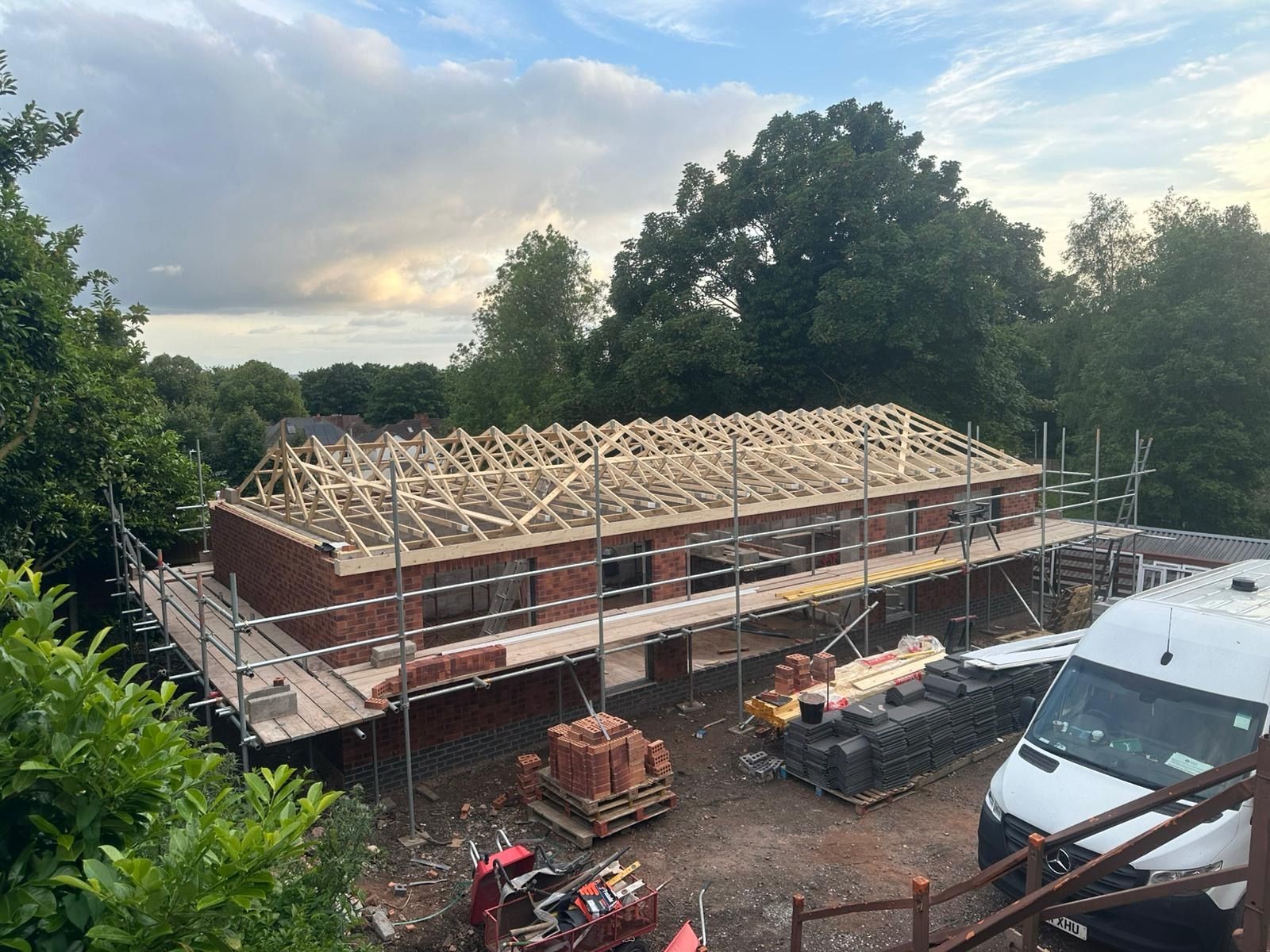 Construction site with a brick building and wooden roof frame surrounded by scaffolding and materials.