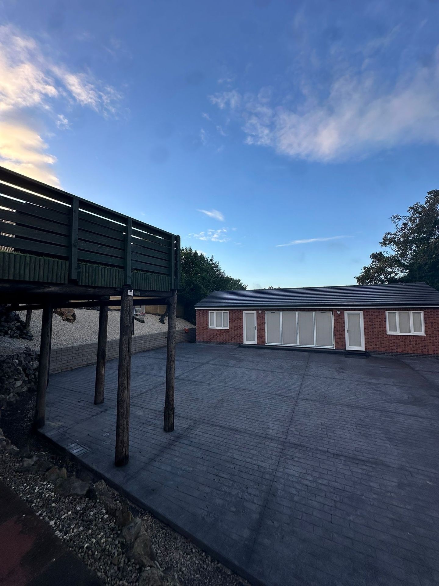 Wide paved area with wooden deck above and a brick building with white doors under a blue sky.